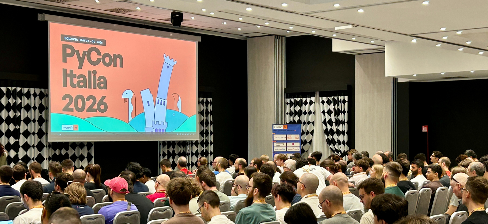 Sala gremita all'apertura di PyCon Italia 2026 a Bologna, sullo schermo la slide con le date della conferenza.