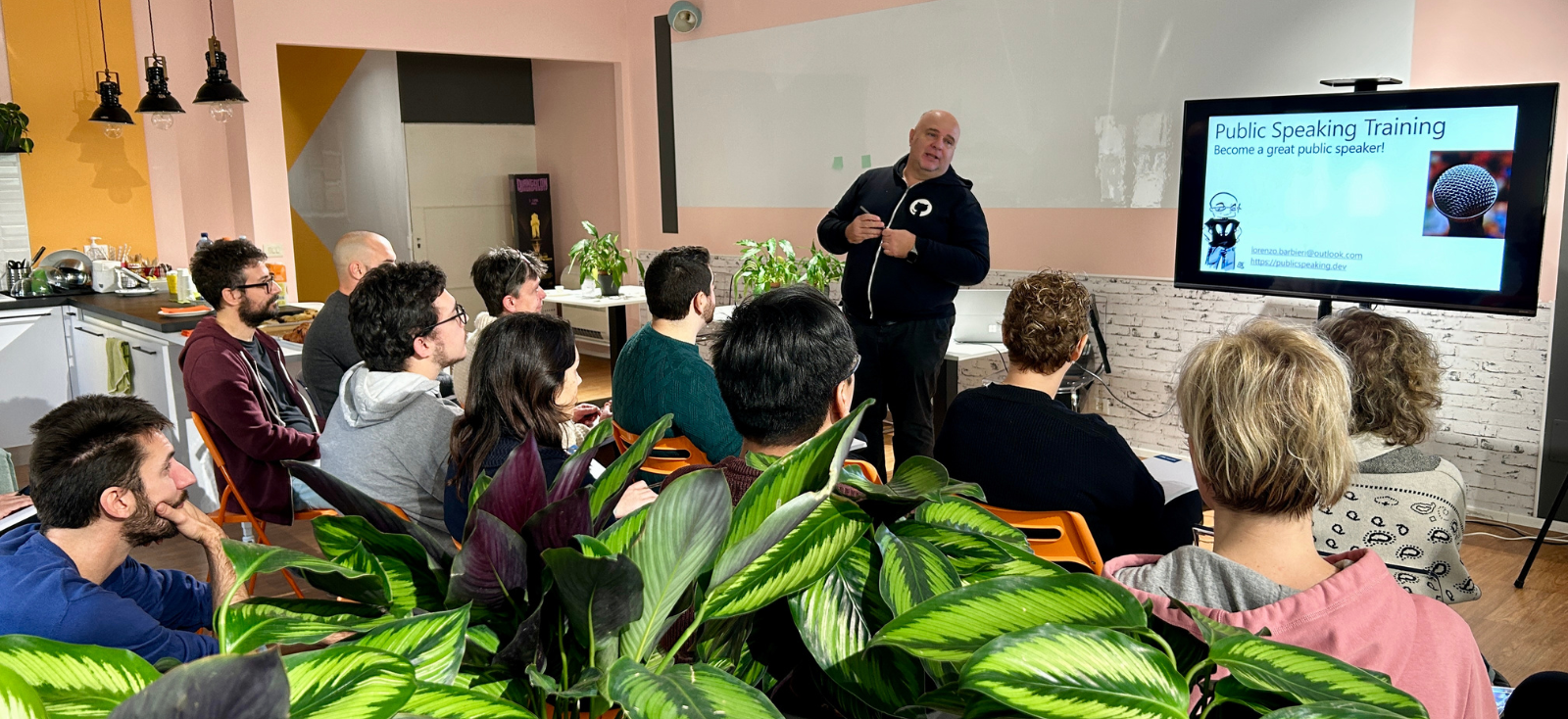 Un team di persone è seduto di fronte a uno schermo e a una lavagna e ascolta una persona che sta facendo una presentazione.